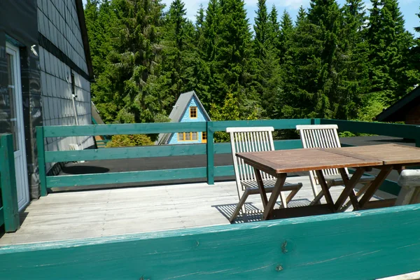 Tisch und Stühle auf dem Balkon am schieferverkleideten Hauptgebäude; dahinter Wald und eine Finnhütte – Fotoarchiv 2013.
