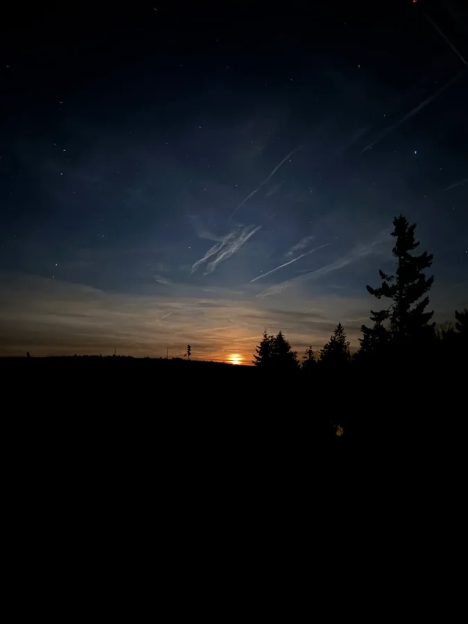 Dunkler Himmel mit Sternen und hellen Wolkenstreifen über einer Waldsilhouette und einem warm leuchtenden Horizont.