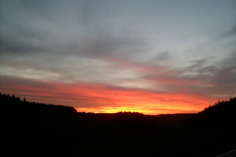 Leuchtend orange und rote Wolken über einer dunklen Waldsilhouette.