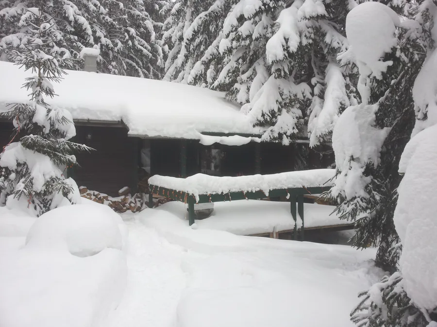 Dunkle Holzhütte und Sitzplatz unter hohen Schneepolstern, eingerahmt von verschneiten Nadelbäumen.